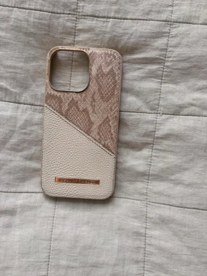iPhone 13 PRO, Beige mobilskal med ormmönster från Ideal of Sweden - Snyggt mobilskal i beige och ljusbrunt med ormmönstrad ovandel och strukturmönstrad nederdel från Ideal of Sweden. Skalet har skador på sidan samt att den gått av i ena hörnet av kameran.