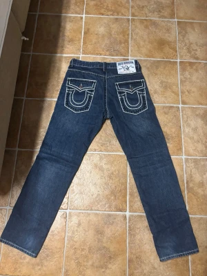 True Religion Jeans  - Kan vara att de är W32 L31 typ Säljer ett par Trues blå jeans med raka ben och tydliga vita kontrastsömmar. Jeansen har stora bakfickor med unik brodyr och patch på linningen. Tillverkade i denim, perfekta för en avslappnad och trendig look.