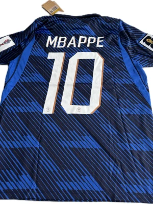 Frankrike VM Mbappé #10 - Snygg blå fotbollströja från Nike med Mbappé och nummer 10 på ryggen. Tröjan har ett grafiskt mönster i olika blå nyanser, vita detaljer och officiella FIFA-märken på ärmarna. Klassisk vit krage och broderad tupp på bröstet. Perfekt för dig som älskar landslagsfotboll.