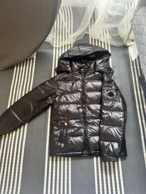 Monclear jacka - Snygg svart monclear dunjacka med glansig finish och huva. Jackan har dragkedja framtill, två fickor med knapp och en extra ficka på ärmen. Perfekt för kalla vinterdagar och ger en modern look. Jackan är köpt på tiktokshop för 1000kr, den har 2 skador som syns på bilderna, en i armhålan som är syd så det inte syns lika mycket och en bak vid nacken som är snyggt lagad med en laglapp. Passar sig som är 12-14 år/ 160-170cm lång