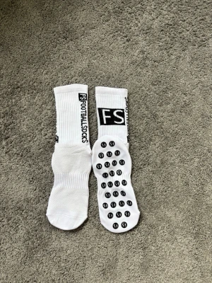 Vita FS Football Socks med grepp - Säljer ett par vita FS Football Socks med svart text längs sidan och grepp på undersidan. Strumporna är ribbade upptill och har en sportig look, perfekta för fotboll eller träning.