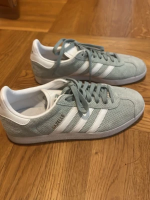 Adidas Gazelle ljusturkosa sneakers - Säljer ett par Adidas Gazelle sneakers i ljusblå mocka med vita detaljer och klassiska tre ränder på sidan. Skorna har vita snören, vit sula och en låg profil. Perfekta för dig som gillar stilrena och sportiga sneakers. Använda en gång!