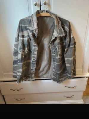 Camo jeansjacka  - Säljer en camouflagemönstrad jeansjacka med råa, slitna kanter och två bröstfickor. Jackan har en avslappnad passform och coola detaljer som ger en edgy look. Perfekt för dig som vill sticka ut med streetstyle-vibbar.