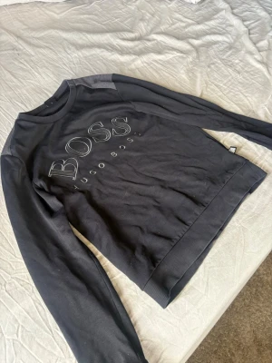 Svart sweatshirt från Hugo Boss - Snygg svart sweatshirt från Hugo Boss med stort BOSS-tryck på bröstet och diskret Hugo Boss-text under. Tröjan har rund halsringning, ribbade muddar och detaljer i mörkgrått vid axlarna. Perfekt för en stilren och avslappnad look.