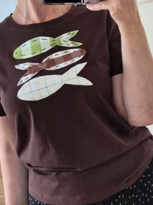 Brun t-shirt med fiskmotiv - Brun t-shirt i bomull med tre applikationer av fiskar i grönt, brunt och vitt med rutigt mönster på bröstet. T-shirten har rund hals och korta ärmar. Perfekt för dig som gillar unika detaljer och lekfull design.