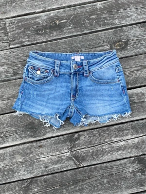 Blå jeansshorts med fransig kant - Säljer ett par blå jeansshorts med fransig rå kant och slitningar. Shortsen har fem fickor, bälteshällor och röda sömmar på bakfickorna. Perfekta för varma dagar och ger en avslappnad vibe.