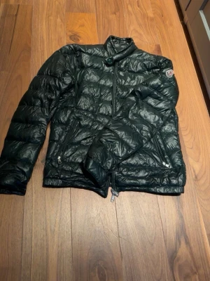 Mörkgrön dunjacka från Moncler - Snygg mörkgrön dunjacka från Moncler med hög krage och diskret logotyp på ärmen. Jackan har två dragkedjeförsedda fickor framtill och quiltad design som ger en modern look. Perfekt för kalla vinterdagar och riktigt lätt att bära.