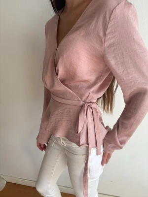 Rosa omlottblus med knytband - Snygg rosa omlottblus med långa ärmar och knytband i midjan. Blusen har en lätt glansig yta och feminin v-ringning. Perfekt att styla med jeans eller kjol för en trendig look.