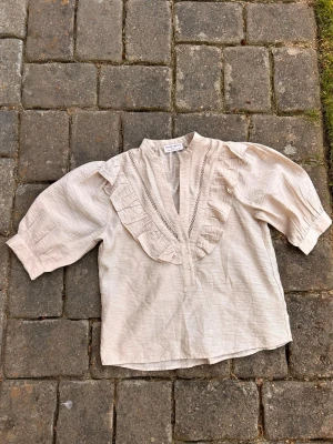 Beige blus med volang och spetsdetaljer - Söt beige blus med trekvartsärm, volang längs den djupa v-ringningen och fina spetsdetaljer framtill. Tillverkad i ett lätt, luftigt tyg som ger en avslappnad men ändå chic look. Perfekt att styla med jeans eller kjol för en trendig outfit. Rymlig modell, vid