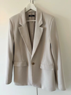 Beige oversized kavaj med knapp - Stilren beige oversized kavaj med en knapp framtill och två fickor med lock. Klassisk krage och lång ärm. Perfekt för en trendig och avslappnad look. Tillverkad i ett mjukt tyg som ger en snygg passform.