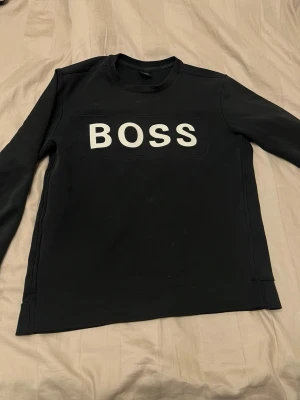 Svart sweatshirt från Hugo Boss - Svart sweatshirt från Hugo Boss med stor vit BOSS-logga på bröstet. Tröjan har rund halsringning och ribbade muddar vid ärmslut och nederkant. Perfekt för en clean och stilren look.