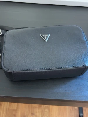 Svart axelväska från Guess - Snygg svart axelväska från Guess med rektangulär form och metallogo framtill. Väskan har justerbar axelrem och dragkedja. Insidan är fodrad med Guess-mönster och har innerfack med dragkedja. Tillverkad i slitstarkt material.