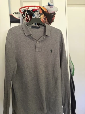 Grå långärmad piké från Polo Ralph Lauren - Klassisk grå långärmad pikétröja från Polo Ralph Lauren med krage, knappar vid halsen och den ikoniska broderade loggan på bröstet. Perfekt till jeans eller chinos för en clean och stilren look.