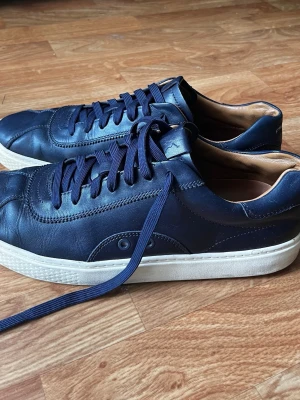 Mörkblå sneakers med vita sula - Snygga mörkblå sneakers i läder med matchande blå skosnören och vit sula. Insidan är fodrad i beige och designen är stilren med diskreta sömmar. Perfekta för dig som gillar en clean look och vill ha något som funkar till det mesta.