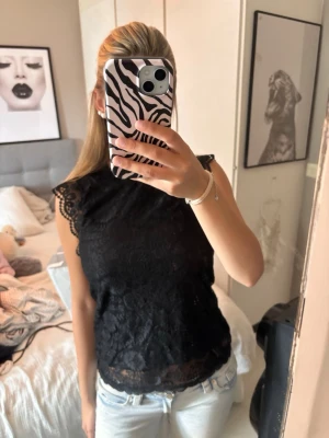 Svart spetsblus med volangdetaljer - Säljer en svart blus i spets med fina volangdetaljer vid axlarna. Toppen är ärmlös och har en hög halsringning. Perfekt att styla med jeans eller kjol för en trendig look.