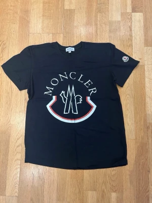 Moncler T-shirt  - Snygg svart t-shirt från Moncler med stor vit och röd logga framtill och en liten patch på ärmen. Klassisk rund halsringning och korta ärmar. Perfekt för dig som gillar streetwear och vill ha något stilrent men ändå med attityd.