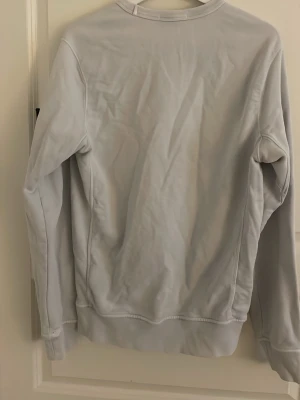 Ljusgrå sweatshirt från Stone Island - Säljer en stilren ljusgrå sweatshirt från Stone Island med klassisk rund halsringning och den ikoniska loggan på vänster ärm. Tröjan har ribbade muddar vid ärmslut och nederkant, perfekt för en clean och avslappnad look.