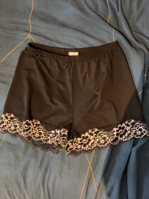 Svarta pyjamasshorts med spetskant - Snygga svarta pyjamasshorts med bred elastisk midja och dekorativ spetskant i blommigt mönster längst ner. Materialet ser mjukt och följsamt ut, perfekt för chillkvällar hemma. Spetsdetaljerna ger en extra touch av stil.