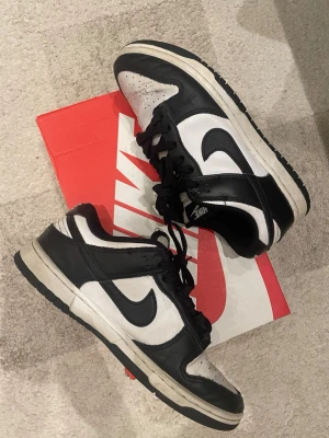 Nike Dunk Low svart/vit sneaker - Säljer ett par Nike Dunk Low sneakers i svart och vitt. Skorna har klassisk låg siluett, svart swoosh och detaljer i läder. Yttersulan är vit med svart kant och skorna har svarta skosnören. Perfekta för dig som gillar streetwear och tidlös design.