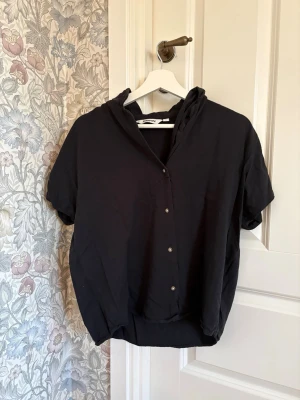 Svart kortärmad blus från Kappahl - Svart blus från Kappahl med korta ärmar och knappar framtill. Blusen har en avslappnad passform och en liten krage som ger en cool touch. Perfekt att matcha med jeans eller kjol för en enkel men snygg look.