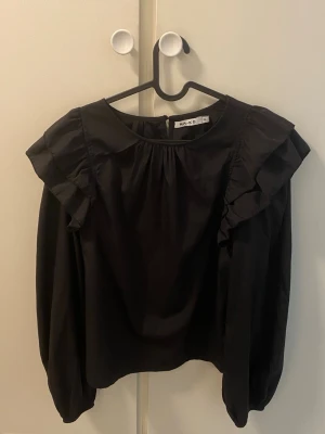 Svart blus med volang från NA-KD - Snygg svart blus från NA-KD med volangdetaljer längs axlarna och rund halsringning. Blusen har långa ärmar med resår vid ärmslut och en lätt glansig finish som ger en elegant look. Perfekt att styla med jeans eller kjol.