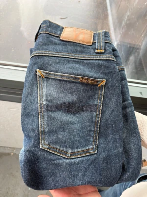 Mörkblå  jeans från Nudie Jeans - Säljer ett par mörkblå jeans från Nudie Jeans med smal passform och klassiska kontrastsömmar. Jeansen har fem fickor, läderpatch bak i midjan och subtila slitningar framtill. Perfekta för dig som gillar en clean och tidlös look.