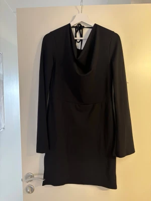 Svart långärmad kortklänning - Stilren svart kortklänning med långa ärmar och snygg draperad urringning. Klänningen har knyt i nacken och en enkel, elegant siluett som passar perfekt till olika tillfällen. Materialet känns mjukt och följsamt.
