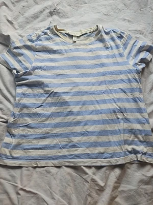 Randig ljusblå och vit t-shirt - En klassisk t-shirt med ljusblå och vita horisontella ränder. T-shirten har rund halsringning och korta ärmar. Tillverkad i mjuk bomull som känns skön mot huden. Perfekt för en avslappnad och enkel stil.