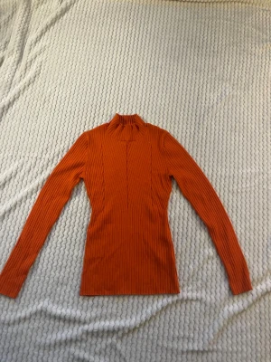 Rostorange ribbad polotopp - Säljer en slim, ribbad polotopp i en snygg rostorange färg. Toppen har lång ärm och hög krage, perfekt för att lyfta din outfit med en poppig färg. Passar dig som gillar trendiga och bekväma plagg.