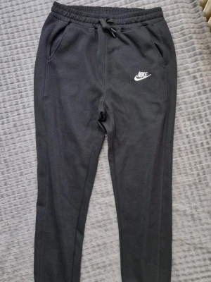 Svarta mjukisbyxor från Nike - Svarta mjukisbyxor från Nike med vit broderad logga på vänster lår. Byxorna har dragsko i midjan och två sidofickor. Tillverkade i mjukt och bekvämt material, perfekta för chill eller träning.