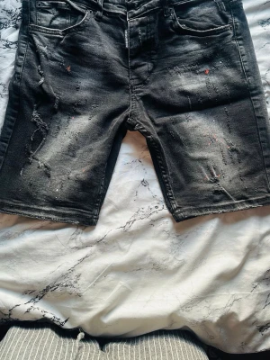 Svarta distressed shorts från Dsquared2 - Säljer ett par svarta jeansshorts från Dsquared2 med distressed detaljer och färgstänk. Perfekta för dig som gillar en edgy och avslappnad stil. Har ett par till vita