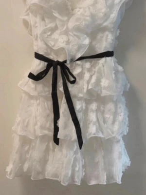 Vit volangklänning med svart knytband - Supersöt vit kortklänning med volanger och blommiga detaljer. Klänningen har v-ringning, korta ärmar och ett svart knytband i midjan som ger en snygg kontrast. Perfekt för sommarens alla tillfällen! Endast använt 1 gång. Pris kan diskuteras💕
