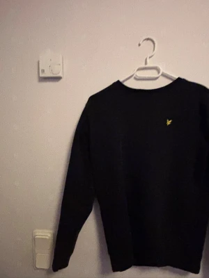 Svart stickad tröja Lyle & Scott - Svart stickad tröja från Lyle & Scott med rund halsringning och den klassiska gula loggan broderad på bröstet. Tröjan har långa ärmar och en enkel, stilren design som passar perfekt till många olika outfits.