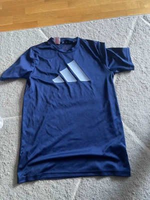Blå Adidas t-shirt med stor logga - Snygg marinblå t-shirt från Adidas med stor vit logga på bröstet. Tillverkad i mjuk polyester som känns skön mot huden. Klassisk rund halsringning och korta ärmar. Perfekt för dig som gillar sportig stil.