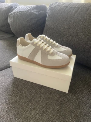 Maison Margiela gats - Snygga sneakers i vitt och beige med detaljer i mocka och klassisk gummisula. Skorna har vita snören och en stilren design som passar till det mesta. Perfekta för dig som gillar minimalistisk och clean stil.