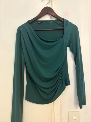 Grön draperad långärmad topp - Snygg grön topp med draperad front och lång ärm. Toppen har en unik asymmetrisk ringning och sitter figurnära. Perfekt för dig som vill ha något stilrent men ändå med en twist.