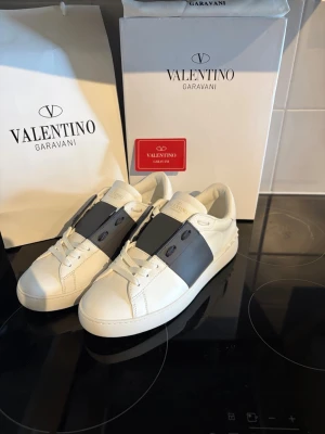 Valentino Garavani  - Snygga vita sneakers från Valentino Garavani med bred svart rem över mitten och diskreta nitar på sidorna. Skorna har klassisk låg profil, rund tå och är tillverkade i slätt läder. Perfekta för dig som gillar stilrena och exklusiva detaljer.