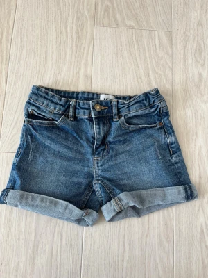 Blå denimshorts med uppvik - Klassiska blå jeansshorts med uppvikta benslut och femficksmodell. Shortsen har normal passform, bälteshällor och stängs med knapp och dragkedja. Perfekta för varma dagar och enkla att matcha med olika stilar. Ena resårbandet trasigt. 