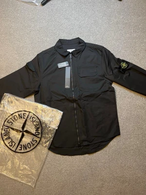 Svart overshirt från Stone Island - Snygg svart overshirt från Stone Island med dragkedja framtill, bröstficka och klassisk patch med logga på vänster ärm. Overshirten har en stilren krage och är tillverkad i ett lätt, slitstarkt material som passar perfekt till lager-på-lager.