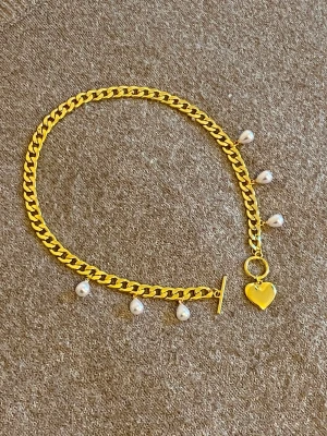 Guldhalsband med pärlor och hjärthänge - Snyggt halsband i guldfärgad metall med droppformade pärlor längs kedjan och ett hjärtformat hänge. Halsbandet har en chunky kedja och stängs med en toggle-lås. Perfekt för dig som vill ha något som sticker ut och känns trendigt.