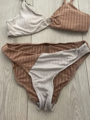 Randig bikini i beige och vit - Säljer en snygg bikini med randigt mönster i beige och vitt. Överdelen har en twistdetalj framtill och smala axelband. Bikinitrosan har samma färgblock och mönster. Perfekt för stranden eller poolen i sommar!