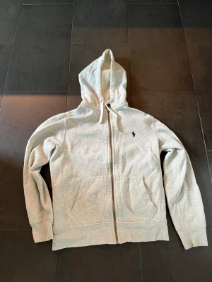 Ljusgrå hoodie från Polo Ralph Lauren - Ljusgrå hoodie med dragkedja från Polo Ralph Lauren. Klassisk design med huva och dragsko, två stora fickor fram och den ikoniska lilla loggan broderad på bröstet. Tillverkad i mjukt bomullsmaterial som är skönt mot huden.
