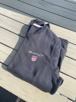 Mörkblå sweatshirt från GANT - Säljer en mörkblå sweatshirt från GANT med klassisk rund halsringning och ribbade muddar. Tröjan har GANT-logga och ett broderat sköldmärke på bröstet. Tillverkad i mjukt material som känns skönt mot huden. Perfekt för en avslappnad och stilren look. Hör av dig vid frågor. Billigare pris vid snabb affär!