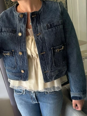 Mörkblå croppad jeansjacka - Trendig mörkblå jeansjacka med rund halsringning och guldiga knappar. Jackan har fyra fickor framtill med dekorativa detaljer och en rak, croppad passform. Perfekt att styla till både klänning och jeans för en cool look! Aldrig använd, prislappen sitter kvar. Nypris 900 kr, säljer för 600 🔥