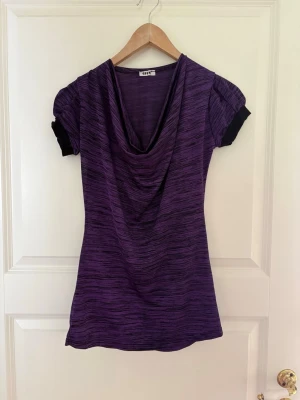 Lila topp med draperad hals - Snygg lila topp med svarta detaljer på ärmarna och draperad halsringning. Toppen har korta ärmar och ett diskret randigt mönster i tyget. Perfekt för dig som vill ha en unik och bekväm stil. Kolla gärna mina andra annonser❗️