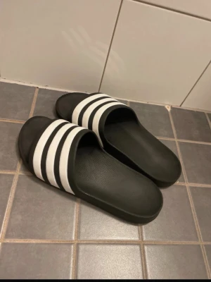 Svarta slides med vita ränder - Säljer ett par klassiska svarta tofflor med tre vita ränder över foten. Tofflorna har en enkel slip-on design och är tillverkade i syntetmaterial, perfekta för chill dagar hemma eller till och från träningen.
