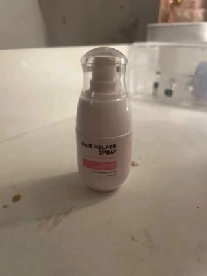 Hair Helper Spray flaska - En  'Hair Helper Spray'. Flaskan har en genomskinlig plastkork och rosa detaljer på etiketten. Perfekt för styling eller vård av håret. Enkel att ta med i väskan.