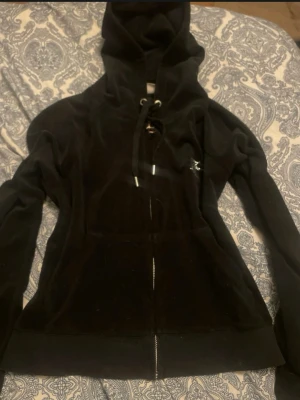 Svart Juicy Couture velourhoodie - Säljer en svart velourhoodie från Juicy Couture med dragkedja framtill, huva med snörning och glittrande Juicy-tryck på ryggen. Perfekt för en chill och trendig look. Klassisk passform och mjukt material.