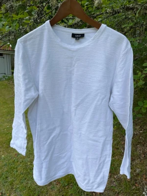Vit långärmad tröja från SMOG - Vit långärmad tröja från SMOG i regular fit. Tröjan har en enkel och stilren design med rund halsringning och lätt texturerat tyg. Perfekt för en clean och avslappnad look.