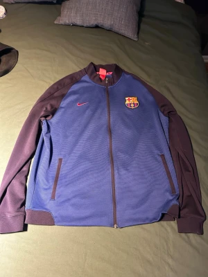 Blå Barcelona träningsjacka Nike - Säljer en blå och vinröd träningsjacka från Nike med FC Barcelona-logga på bröstet. Jackan har hel dragkedja, två fickor framtill och ribbade muddar. Perfekt för dig som älskar fotboll och vill representera ditt lag. Köpt i Spanien 2016 på Barca shoppen